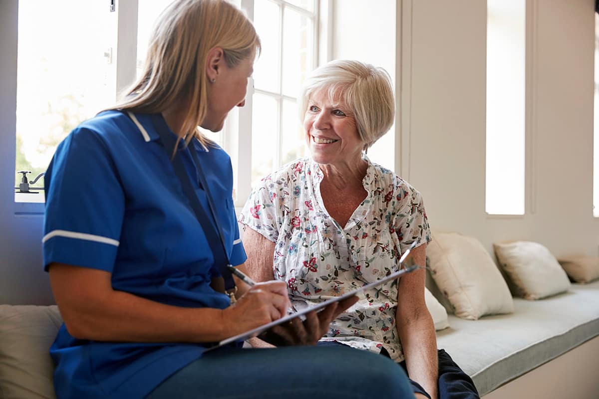 Home care nurse sits on a couch with a woman while chatting and taking notes